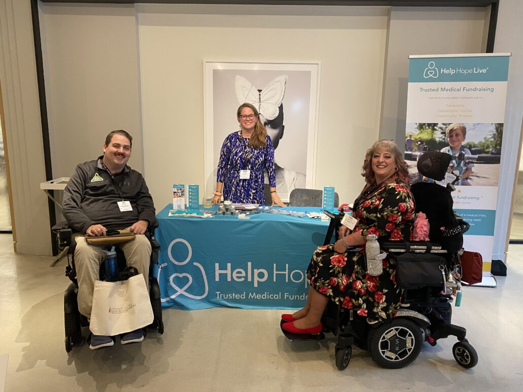 Sonny has a flowered blue dress, light skin, sandy brown hair past her shoulders, light skin, and black-framed glasses as she stands behind a Help Hope Live teal table at Reeve Summit. Two individuals in black power chairs are in front of the table, including client and ambassador Ian Burkhart.