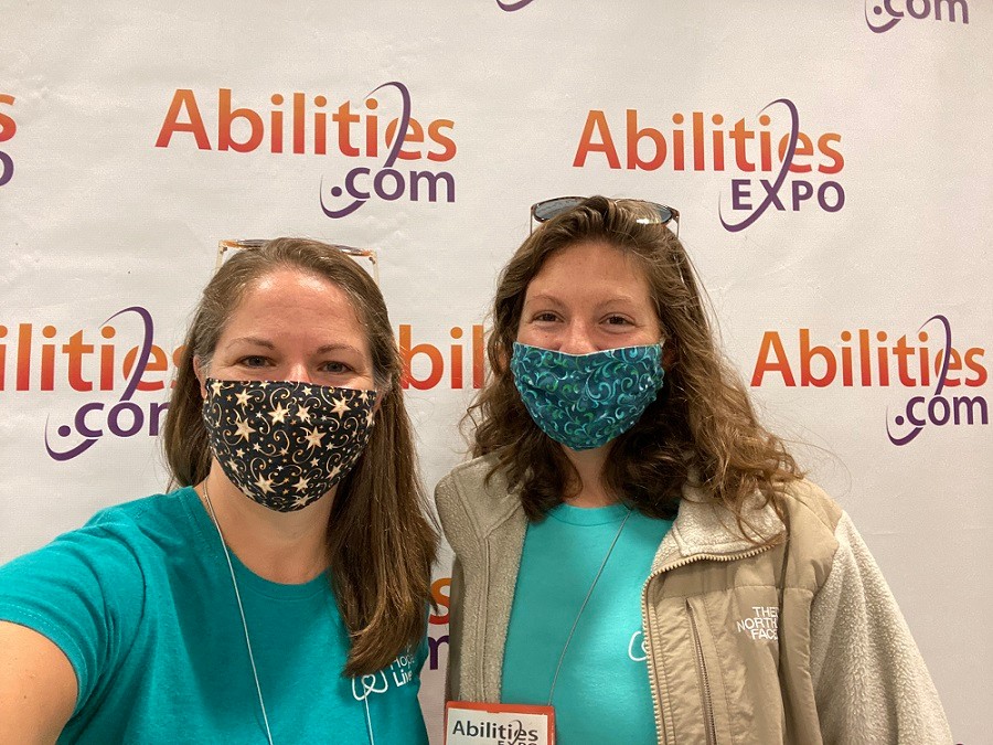 Kate and Sonny from Help Hope Live wear teal Help Hope Live t-shirts and colorful face masks as they pose in front of a Abilities Expo step-and-repeat backdrop that reads Abilities.com.