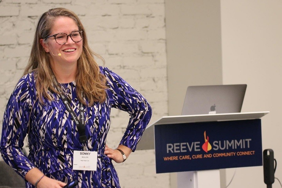 Sonny smiles as she presents at the Reeve Summit. She wears a dark blue printed dress and has light skin, a microphone near her mouth, glasses, and sandy hair past her shoulders.