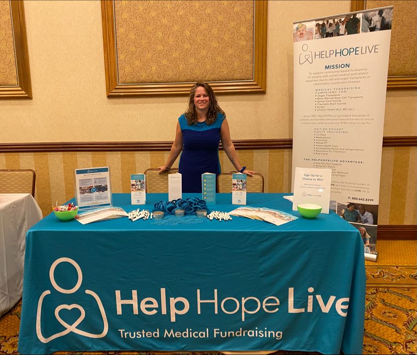 Sonny from Help Hope Live wears a dark blue and light blue colorblocked dress as she stands behind a teal Help Hope Live table at an event.