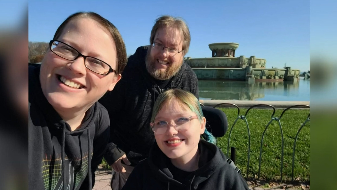 17-year-old Alivianna with her parents is outdoors seated in her black power chair. She has light skin, red lipstick, glasses, and dyed hair in a ponytail.