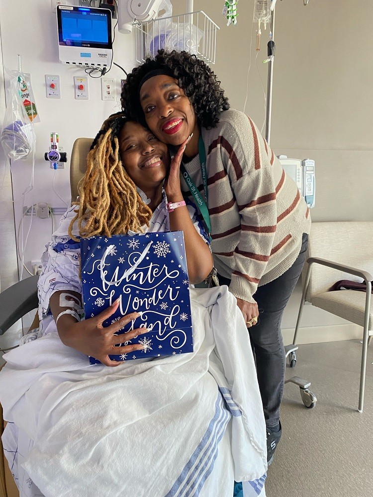 A patient with brown skin and dyed blonde black hair smiles as she holds a Bags of Hope bag that says Winter Wonderland. She is in the hospital wearing a hospital gown and bracelet. Beside her is a medical professional with brown skin and curly brown hair wearing a beige sweater with red stripes.