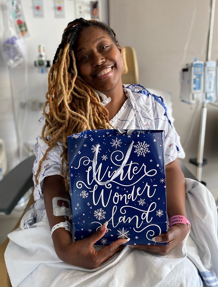 A patient with brown skin and dyed blonde black hair smiles as she holds a Bags of Hope bag that says Winter Wonderland. She is in the hospital wearing a hospital gown and bracelet.