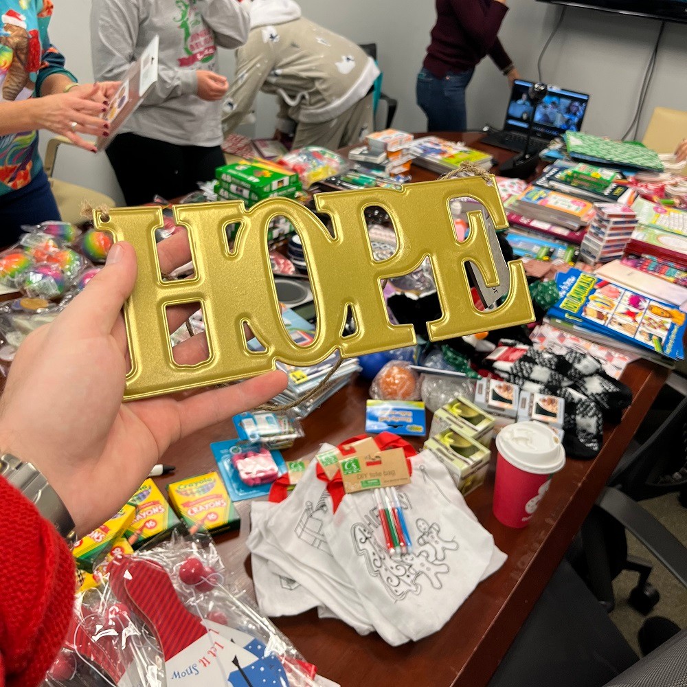 A Help Hope Live team member's hand raises a gold HOPE sign for a photo with a conference table covered in Bags of Hope items like coloring books visible behind the gold sign.