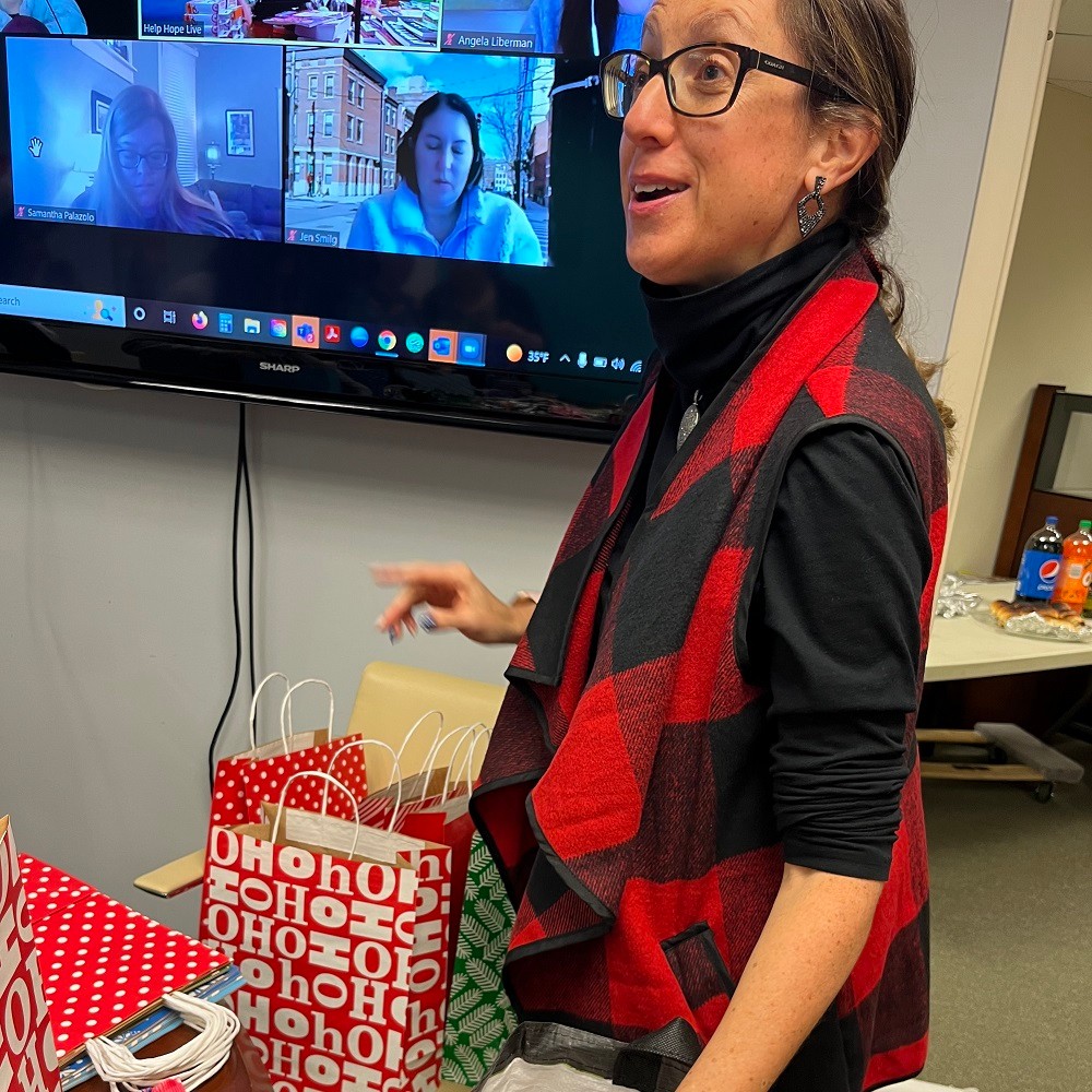 Help Hope Live's Brooke Schostak wears a festive red and black tartan vest as she packs Bags of Hope at the Help Hope Live offices. Several remote members of the Help Hope Live team are visible on a Zoom call on the large television screen behind Brooke.