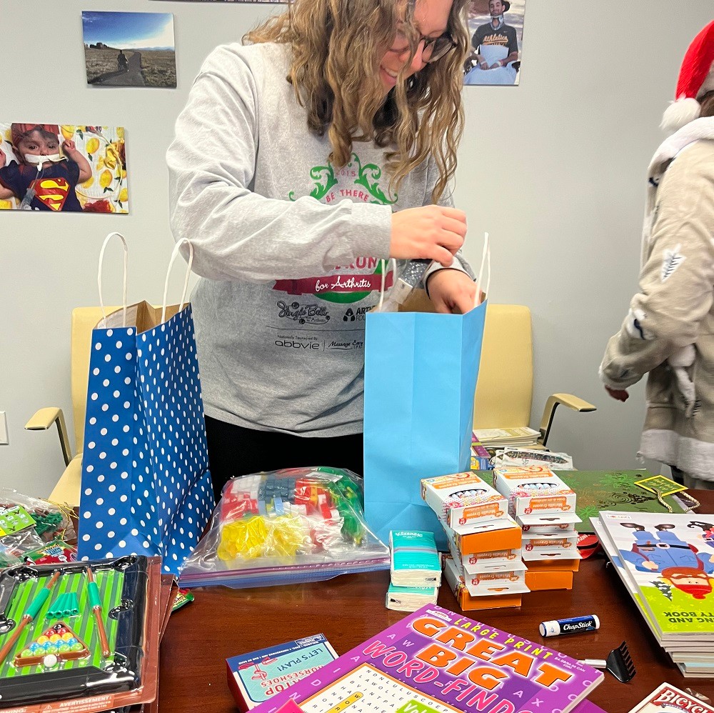 Help Hope Live's Katherine Lacouture wears a festive gray holiday sweater as she packs a Bags of Hope bag with coloring books, puzzle books, crayons, and other items at the Help Hope Live offices.