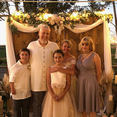 Liver transplant recipient Jeremy is pictured in a wedding setting with light skin, white hair, a white goatee, and a formal white shirt. Near him stand a woman in a white wedding dress and three children in formal garb.