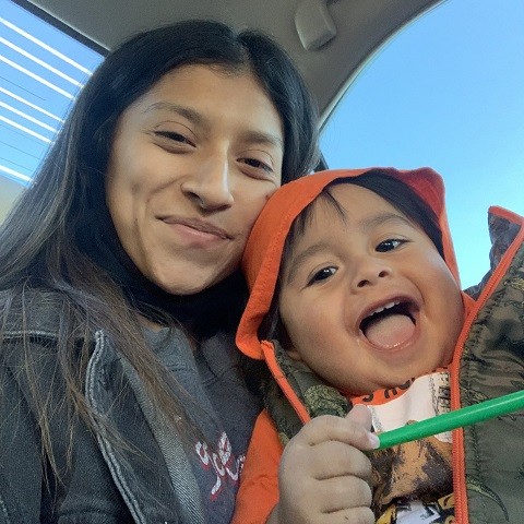 Kidney transplant recipient Jocelyn smiles with her young son on her lap, who wears a bright orange hoodie. Both she and her son have light brown skin, black hair, and dark eyes.