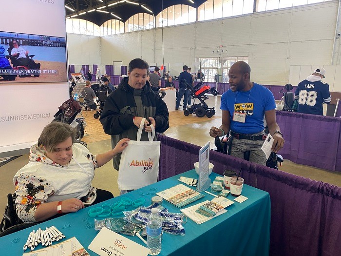 Justin from W.O.W. Wonders on Wheels is strapped into a black upright mobility device paused next to the Help Hope Live teal tablecloth-covered booth at Abilities Expo Dallas. Justin has brown skin and short-cropped black hair and beard. He wears a bright blue W.O.W. t-shirt. He addresses a male-presenting light skinned Expo attendee, standing, and a female-presented light skinned attendee who is seated in a black mobility device as they look at the materials on the Help Hope Live booth table.