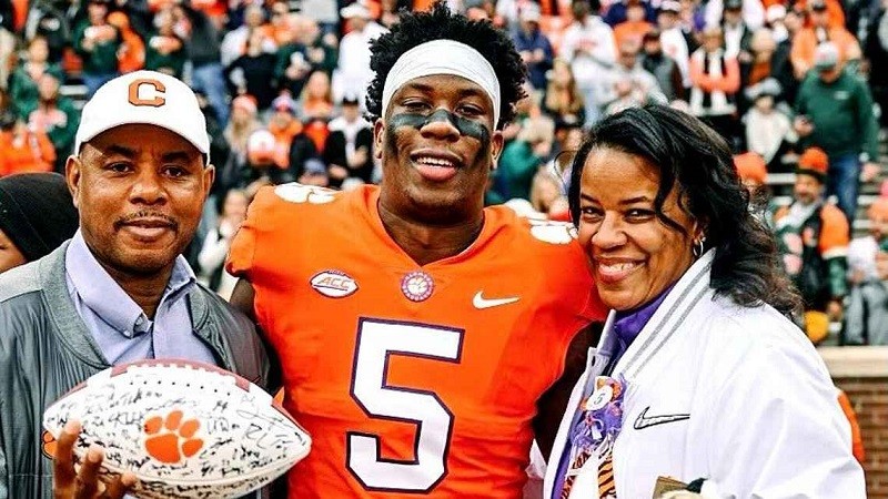 Keith Henry holds a souvenir Clemson football and a Clemson cap as he stands next to his son KJ Henry and his wife. KJ wears a bright orange Clemson football jersey with the number 5 and a white athletic headband across his forehead. Keith and KJ have brown skin - KJ has curly black hair protruding from behind the headband. KJ's mother has brown skin and black hair just past her shoulders.