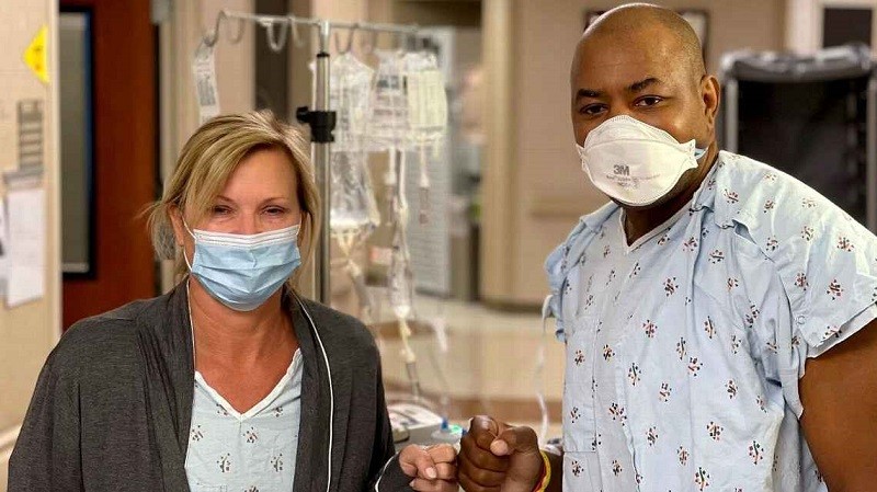 Keith Henry and his kidney donor are wearing hospital gowns and masks in a hospital corridor. Keith's kidney donor Karen Eaten has light skin and blonde hair tied back. Keith Henry has brown skin and a bald head and stands about a foot taller than Karen.