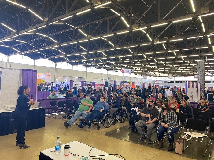 Executive Director Kelly L Green wears a black suit and heeled boots as she addresses a packed crowd of about fifteen Abilities Expo Dallas attendees. She has light skin and dark brown hair in a ponytail.