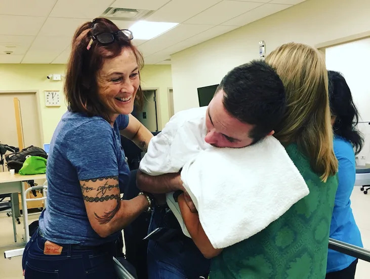 Lisa Nigro cares for her son Nick Nigro. Lisa is smiling wide and has light skin, tattoos on her right arm, and red hair to her shoulders. Nick is 30 years old and has light skin and short black hair, and he leans on a professional in scrubs's shoulder with a supportive towel or cushion around his neck.