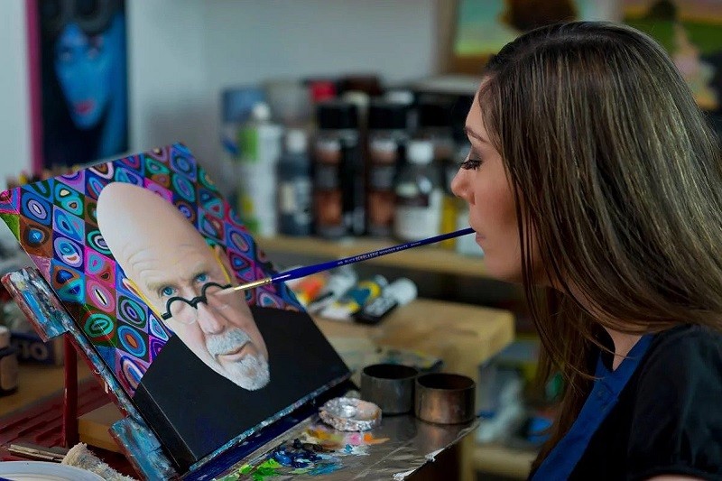 Mariam Pare holds a paintbrush in her mouth as she works on a stunning celebrity portrait on canvas. She has long brown hair and light skin.