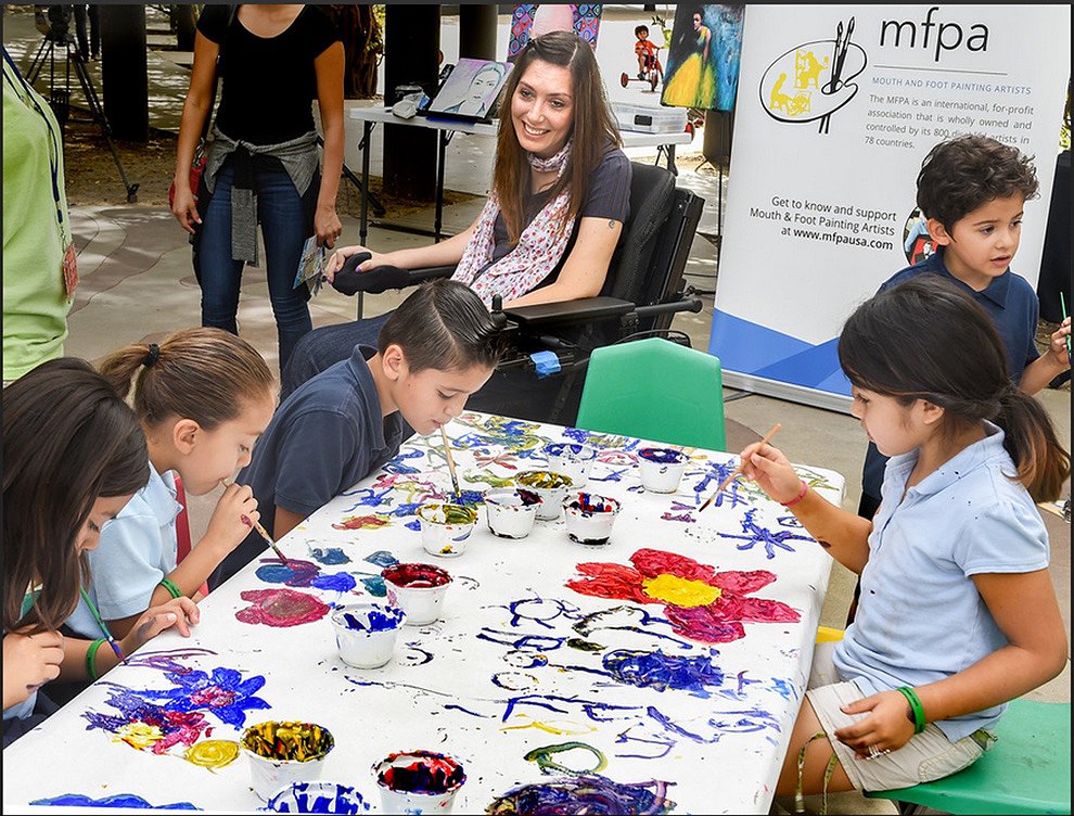 Seated in a black power chair, Mariam grins at a table where five children paint brightly-colored art on a white table using their mouths to paint just like she does. Mariam has long brown hair and light skin.