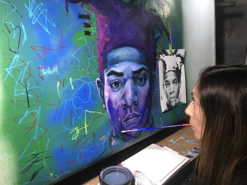 Mariam works on a stunning large-scale portrait of Jean-Michel Basquiat based on a small black and white photograph. The portrait features vivid shades of blue, purple, and green. Mariam holds a paintbrush in her mouth as she works with long brown hair and light skin.