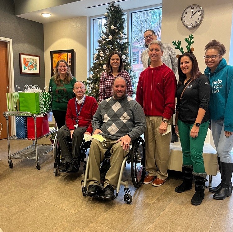 The Help Hope Live team smiles in the lobby of Magee Rehabilitation with board member Ron Siggs and two Magee professionals who are seated in their wheelchairs in front of a Christmas tree.