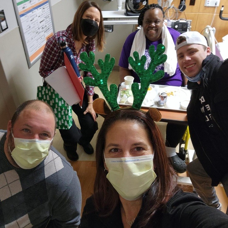 Wearing masks, the Help Hope Live team take a selfie as they deliver a Bags of Hope gift to a patient who is seated in a hospital room. She has brown skin and a purple t-shirt.