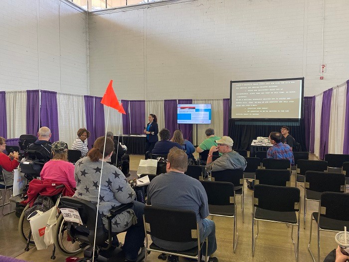 Abilities Expo Dallas attendees watch Executive Director Kelly L Green present about Help Hope Live. Some are seated in black chairs while other are seated in manual or power wheelchairs.