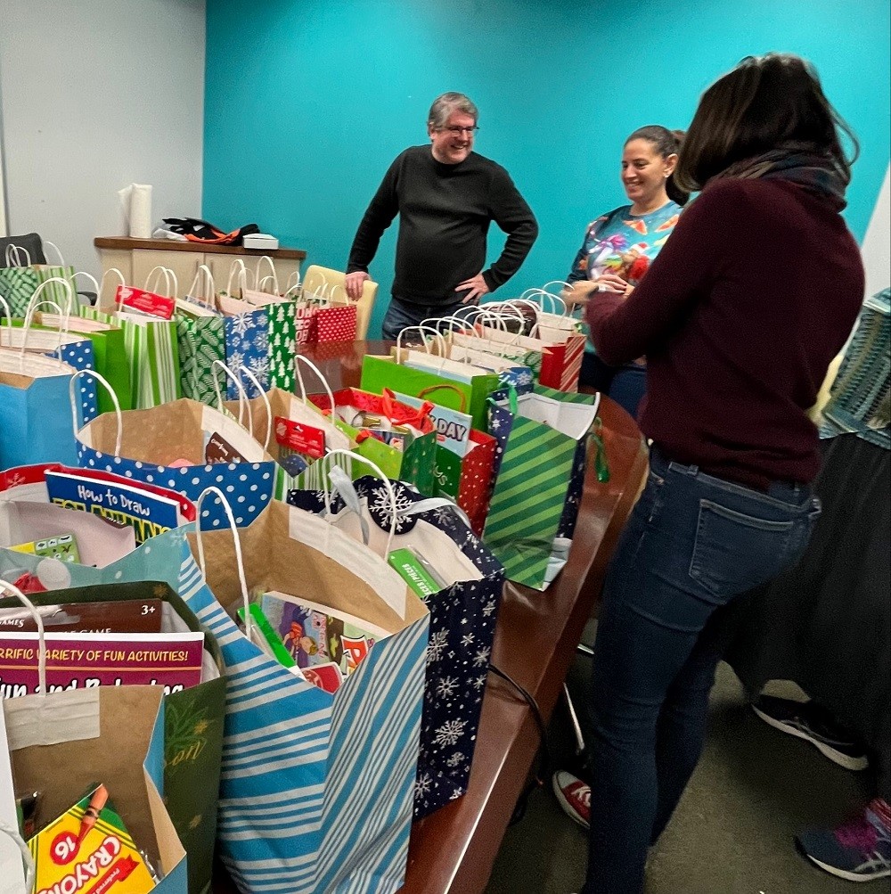 Help Hope Live's David Cahill, Kelly Green, and Shannon Shensky stand near a conference table at the Help Hope Live offices covered in fully packed and brightly-colored Bags of Hope bags.