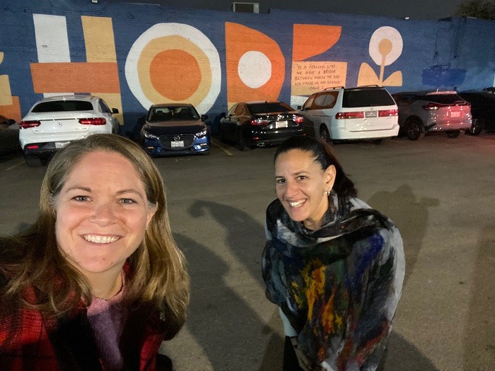 Kelly Green and Sonny Mullen grin as they take a selfie in front of a large orange and white HOPE mural above parked cars outdoors in Dallas.