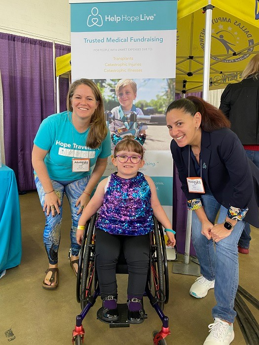 Sonny Mullen and Kelly L Green hunch down slightly to get a photo with client Riley Drury, a seven-year-old in a manual wheelchair. Riley has light skin, purple-framed glasses, and a dazzling sequin-covered blue and purple sleeveless dress shirt.