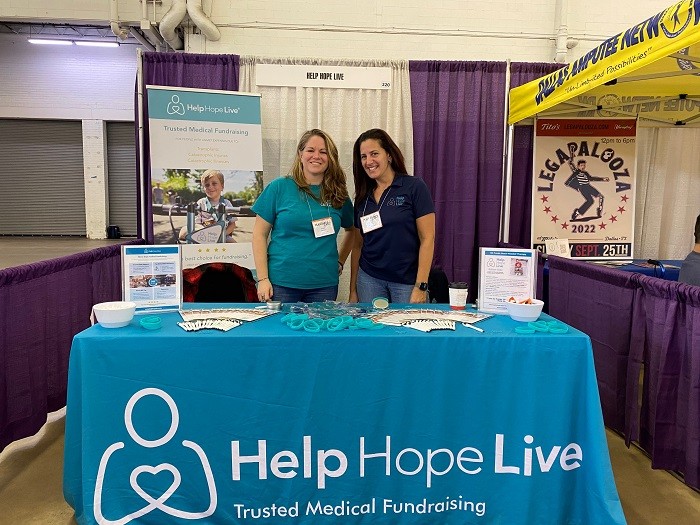 Sonny Mullen and Kelly Green stand at the Help Hope Live booth of Abilities Expo Dallas near a Help Hope Live banner. The table before them is covered in brochures, wristbands, and more over a bright teal tablecloth that reads Help Hope Live Trusted Medical Fundraising.