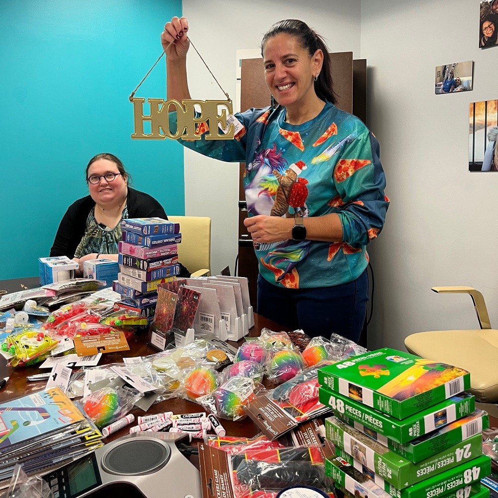 Help Hope Live's Kelly Green and Stacia Moak are in the Help Hope Live offices with a teal wall visible behind them as they assemble Bags of Hope from a stacked conference table of puzzles, knick-knacks, coloring books, and more. Kelly raises a gold HOPE sign and smiles, wearing a brightly-colored festive sweater.