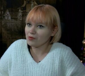 Alivianna Gallup is being interviewed in this screenshot. The 17-year-old has light skin, light blue eyes, and winged eyliner with a shoulder-length dyed bob and a white sweater.