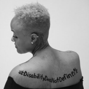 In a dramatic black and white portrait, Keisha Greaves lookes over her left shoulder with her eyes downcast. Painted across her back is #DisabilityDoesNotDefineMe. She has short curly hair, a feather-like tattoo behind her left ear, and a nose stud.