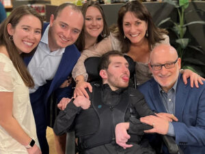 22-year-old Ethan Kadish is strapped int a black harness and is seated in a black wheelchair. He has short curly brown hair, light skin, faint stubble, and a pink tongue protruding from between his lips. Surrounding him are his parents and other family members, dressed in formal attire and smiling.