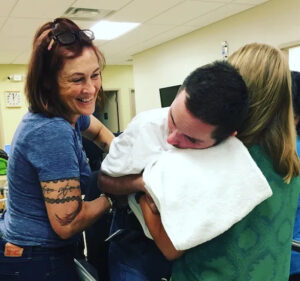 Lisa Nigro cares for her son Nick Nigro. Lisa is smiling wide and has light skin, tattoos on her right arm, and red hair to her shoulders. Nick is 30 years old and has light skin and short black hair, and he leans on a professional in scrubs's shoulder with a supportive towel or cushion around his neck.