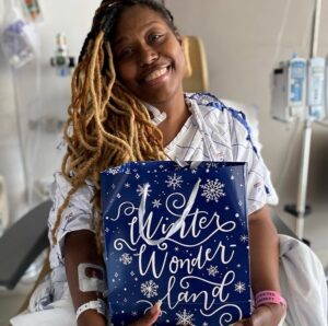 A patient with brown skin and dyed blonde black hair smiles as she holds a Bags of Hope bag that says Winter Wonderland. She is in the hospital wearing a hospital gown and bracelet.