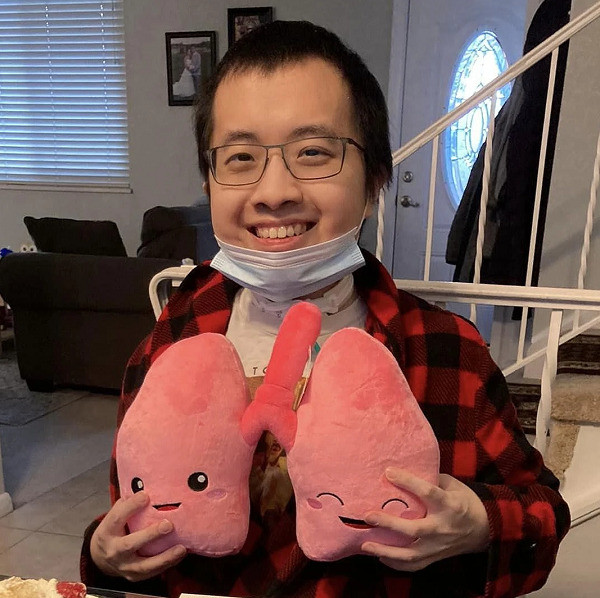 Lung transplant recipient Austin Yang is 26 years old and Asian with black narrow-rimmed glasses and short black hair. He smiles and holds a plushie shaped like a set of lungs.