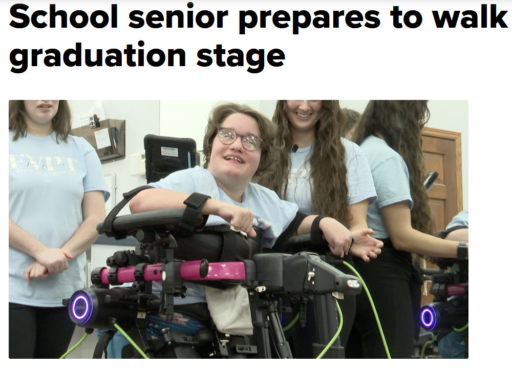 18-year-old Ella Weiske is pictured beaming as she stands in a Trexo robotic device. She wears a light blue t-shirt that matches the shirts of three peers standing near her. She has light skin, short brown hair, and glasses.