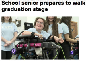 18-year-old Ella Weiske is pictured beaming as she stands in a Trexo robotic device. She wears a light blue t-shirt that matches the shirts of three peers standing near her. She has light skin, short brown hair, and glasses.