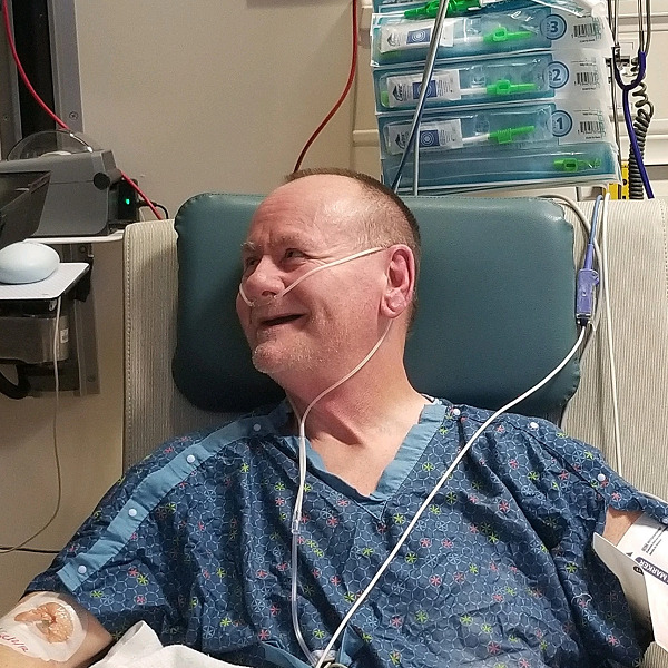 Double lung transplant recipient Greg Fulton is in a hospital setting with a tube connected to his nose. He has light skin and a shaved head and is smiling.