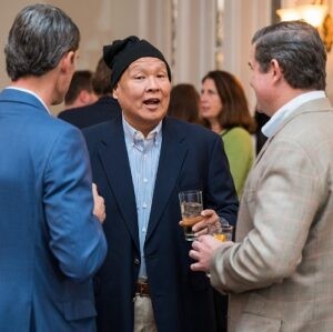 Bruce Ikeda is pictured at the 2022 Live It Up! gala to support Help Hope Live. He is wearing a blue colared shirt and navy jacket as he speaks with two men in similar garb. He is Asian with a black knit cap over his head in contract to his formal attire.