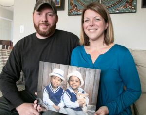 Jason Teagarden's parents hold a photo of their adopted sons. Jason's father has light skin, a shirt brown beard, and a ball cap. Jason's mother has shoulder-length blonde hair and light skin. Jason and his brother are young children in the photo wearing matching navy and white outfits. Both boys have light brown skin.