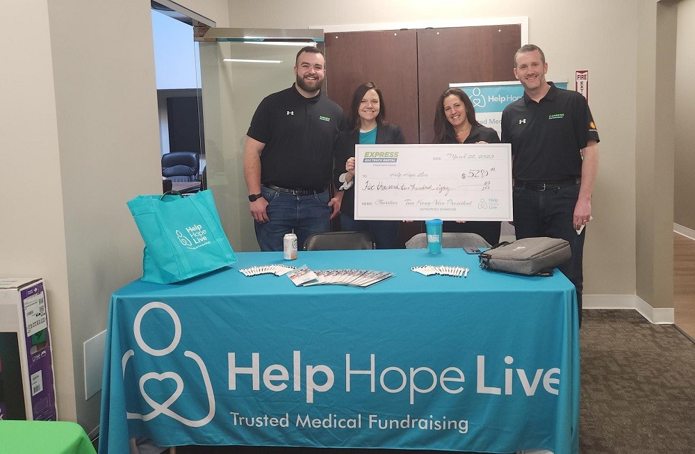 Shannon and Kelly from Help Hope Live hold up a giant check from Express 4x4 Truck Rental for $5,280 made out to Help Hope Live. Two Express staff members stand beside Kelly and Shannon wearing black polo shirts. In front of them is a Help Hope Live booth table with a teal tablecloth, brochures, and pens.