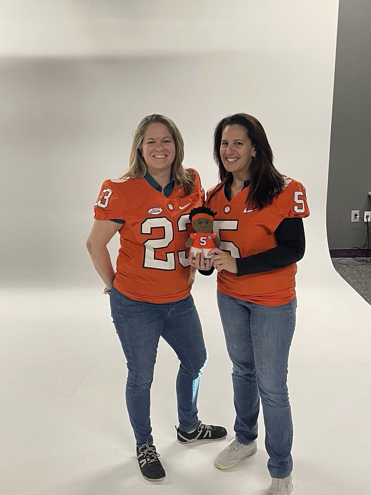 Help Hope Live’s Sonny Mullen and Kelly Green hold a knit doll of KJ Henry, which has brown skin, curly black hair, and an orange #5 Clemson jersey. Kelly wears the same #5 orange Clemson jersey while Sonny wears a #23 orange Clemson jersey.