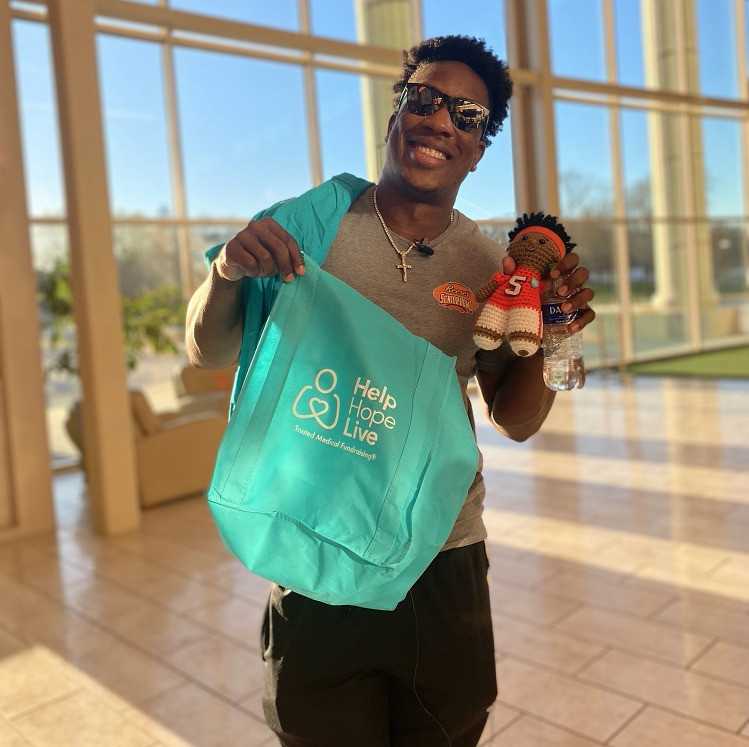 KJ Henry smiles and holds up a teal tote bag that reads Help Hope Live: Trusted Medical Fundraising in one hand and a knit doll of himself in the other hand. The doll has brown skin and a football uniform with a Clemson orange #5 jersey. KJ wears sunglasses and a cross necklace.