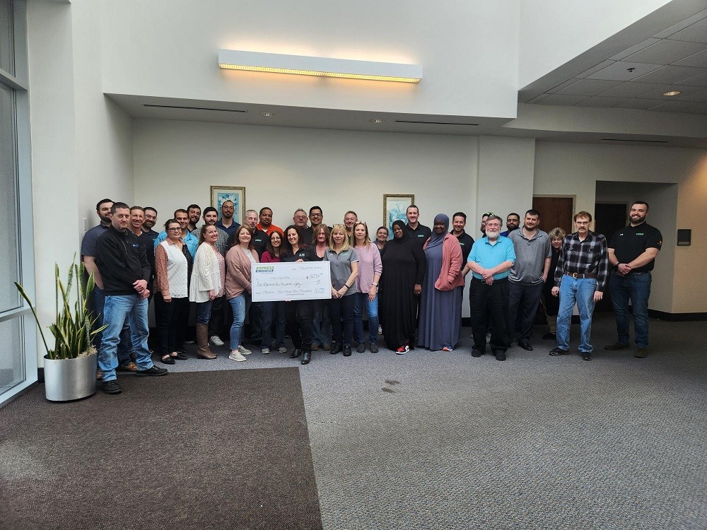 The Express 4x4 Truck Rental Team stands in a building lobby with Help Hope Live’s Kelly Green in the center holding a giant check for $5,280. The Express team is a diverse mix of genders, ages, and ethnicities.