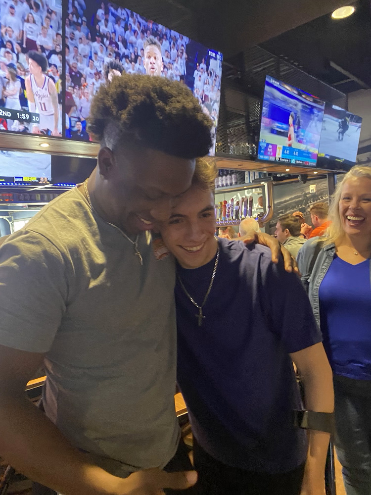 At a sports bar, KJ Henry hugs Austin Somerville. KJ has brown skin and curly black hair and is a head taller than Austin, who has light skin, brown hair, and a cross necklace with braces around his forearms.