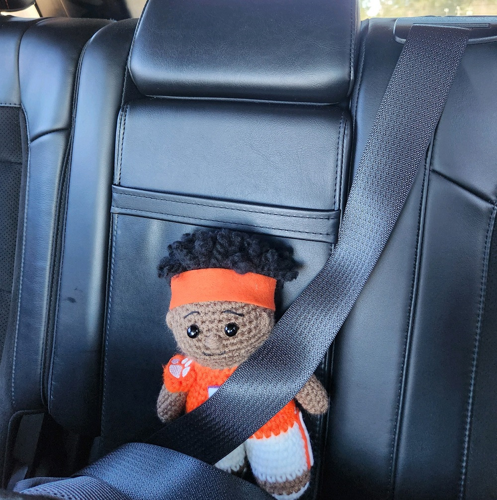 A knitted KJ Henry doll with brown skin and an orange #5 Clemson jersey sits in a car with the seatbelt fastened.