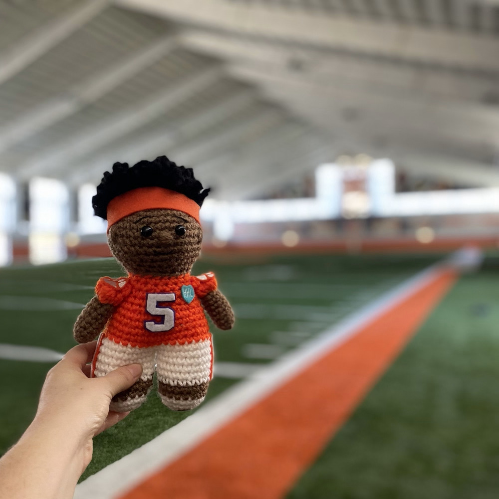 A knitted KJ Henry doll with brown skin and an orange #5 Clemson jersey is at an indoor football practice field.