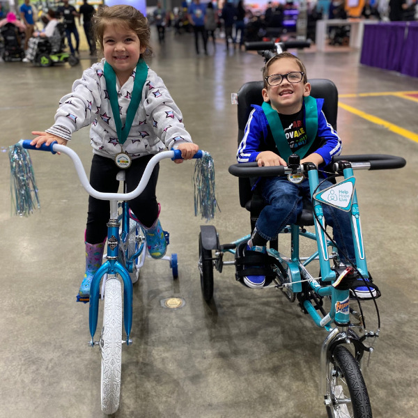 4-year-old Austin and his twin sister sit side-by-side on bikes—his adaptive with three wheels and hers traditional with training wheels—on the expo floor at Abilities Expo LA.