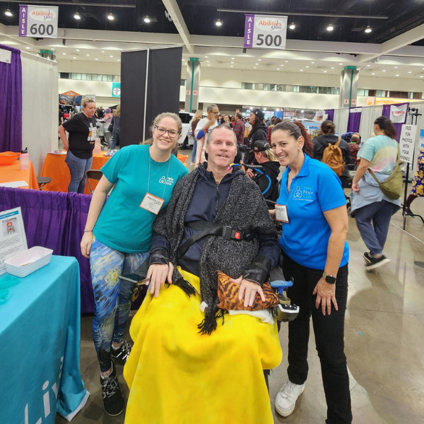 Sonny Mullen and Kelly L Green are at Abilities Expo LA standing on either side of client Gary Suydam, who is seated in his power chair with a warm bright yellow blanket draped over his lap. Gary has light skin and short gray hair.