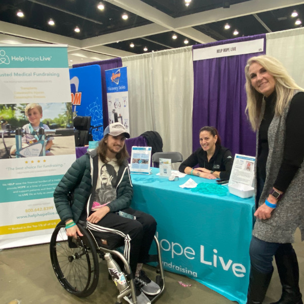 Kelly L Green is seated at the teal tablecloth of Abilities Expo LA's Help Hope Live booth as she meets Spencer Sherwin, who is 26 years old and seated in his black wheelchair, and his mom Meredith, who is standing next to the booth.