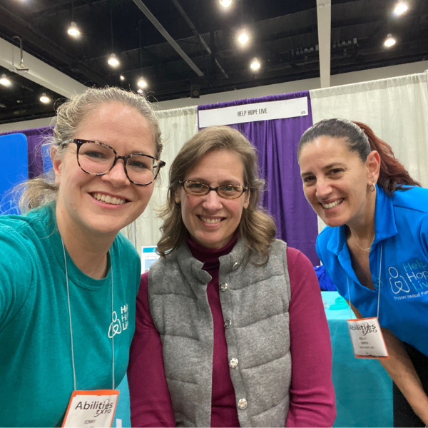 Sonny Mullen and Kelly L Green from Help Hope Live at Abilities Expo Los Angeles meet client Lyena, who has light skin, tortoiseshell glasses, gray eyes, and sandy brown hair. She is seated in her wheelchair.